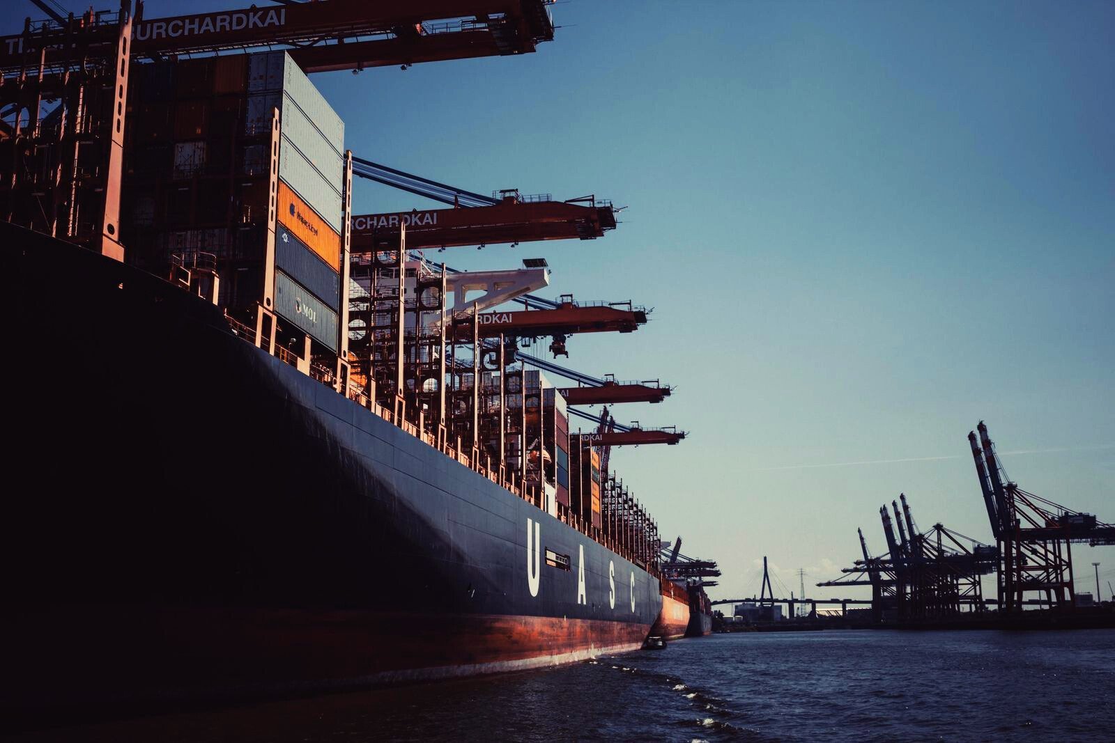 Large cargo ship being loaded with containers at a busy port terminal with multiple cranes under clear blue sky